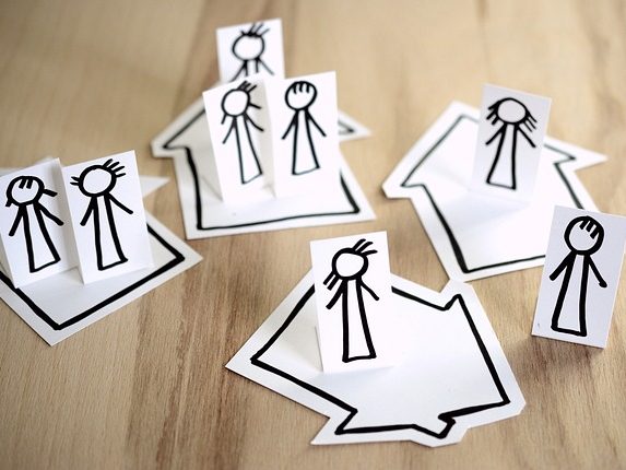 Familie Papierfiguren in verschiedenen Positionen auf Papierhäusern und Pfeilen angeordnet.